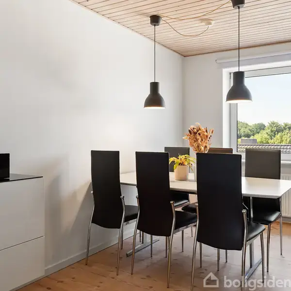 Et elegant spisebord med seks sorte stole står i et lyst rum. Over bordet hænger to sorte lamper. Et vindue viser udsigt til træer udenfor.