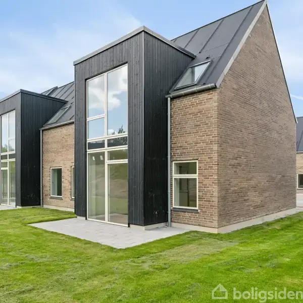 En moderne bygning med stor glasfacade og stejlt skrånende tag stående i et boligområde. Murstensvæg kombineret med sorte paneler. Omgivet af en velplejet græsplæne.