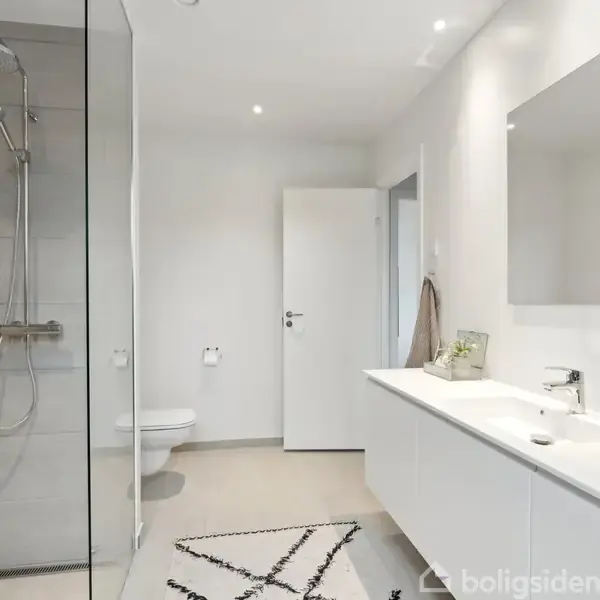 Badeværelse med bruser i glasskabinet, dobbeltvask med spejl over og moderne armaturer. Lyse farver præger rummet med et minimalistisk design. En åben dør fører til et tilstødende rum.