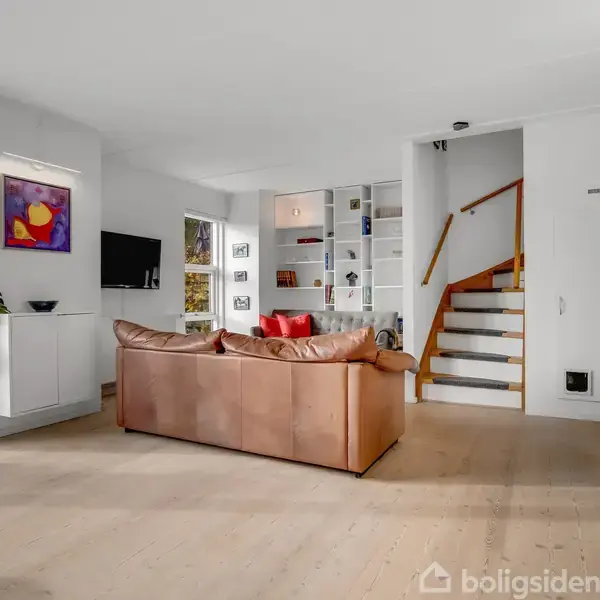 En brun lædersofa står i en lys stue med trægulv, fladskærms-TV, kunstmaleri og en indbygget reol. En trappe fører opad, og en plante står ved en dør.