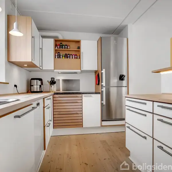 Et moderne køkken med hvide skabe og træbordplader omgiver et rustfrit stål køleskab. På bordet står en kaffemaskine; der er krydderier i en åben hylde. Vinduet lader naturligt lys strømme ind.