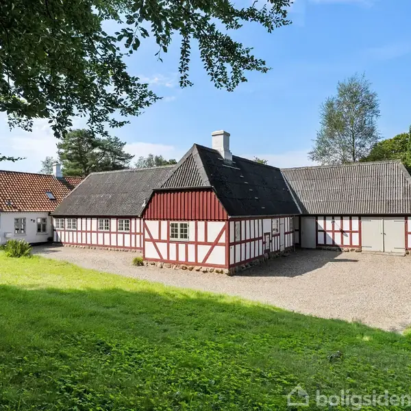 Bindingsværkshus står stille i solen med røde og hvide vægge samt mørkt tag. Grusvej foran bygningen omgivet af grønne marker og træer under en blå himmel.