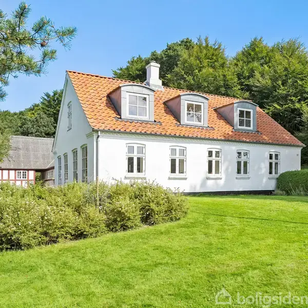 Huset er hvidt med rødt tag og to kviste, beliggende i en frodig have omkranset af træer. I baggrunden ses en anden bygning med røde detaljer.