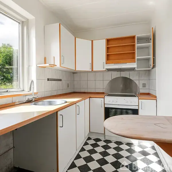 Et lille, lyst køkken med sorte og hvide ternede gulvfliser. Køkkenet har hvide skabe, en indbygget ovn, rund vask og et lille vægmonteret bord under et stort vindue.