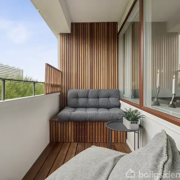 Balkon med træbeklædt bænk og grå hynder, placeret ved siden af et lille bord med en plante. To lejlighedsbygninger ses i baggrunden over træernes toppe.