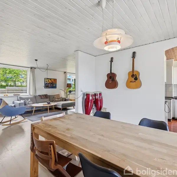 Træbord omgivet af stole, står i spisestue. To guitarer hænger på væggen, trommer ses. Stuen har sofaer ved vindue. Køkkenet i baggrunden ses med hvidt interiør.