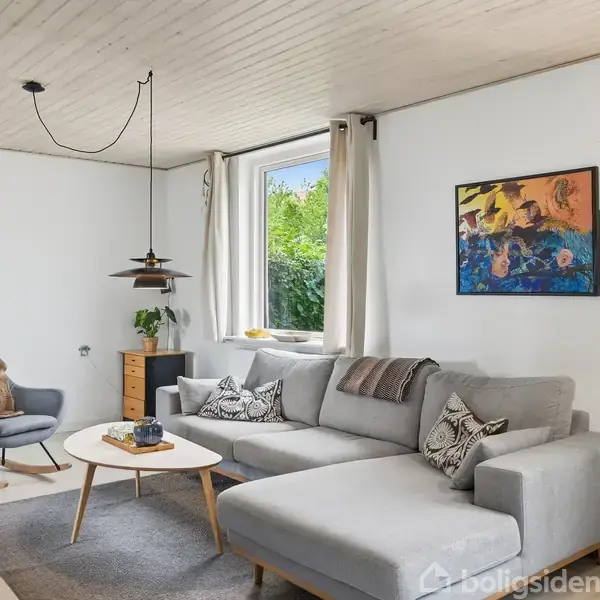 Grå hjørnesofa står i stue med lyskilder fra vindue og gulvlampe, omgivet af hvide vægge og træloft. En gyngestol står ved siden af et lille bord med en plante.