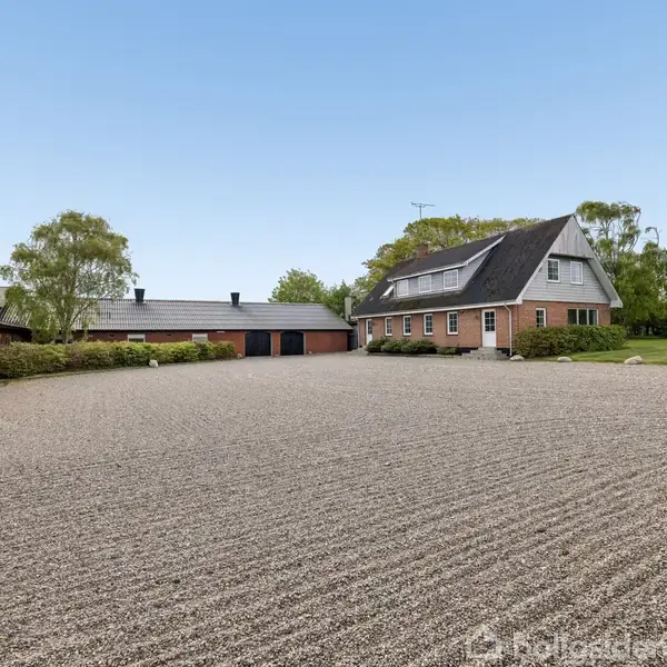 En stor, rød murstensbygning med sort tag omgivet af en grusplads. Til venstre ses en garage og til højre grønne træer og en plæne under en klar, blå himmel.