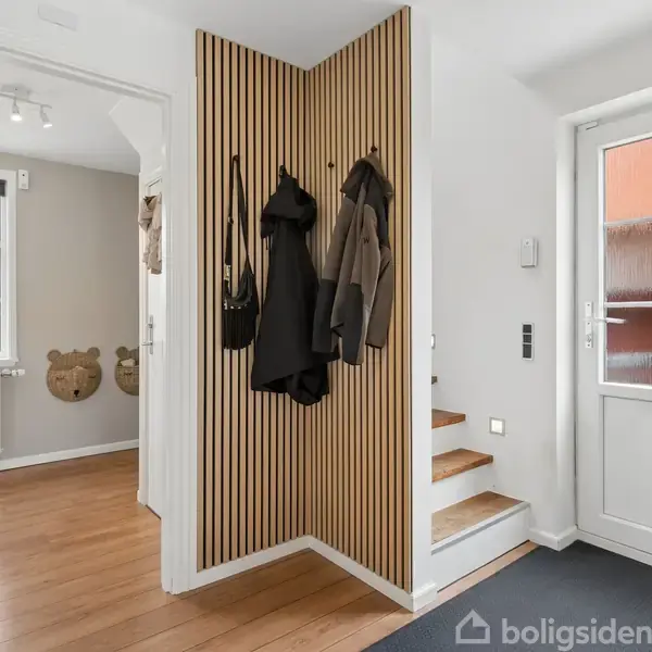 Garderobe med jakker hængende på kroge, placeret i en lys gang med trægulv. Dør til højre fører ud, mens der er et rum med vindue til venstre.