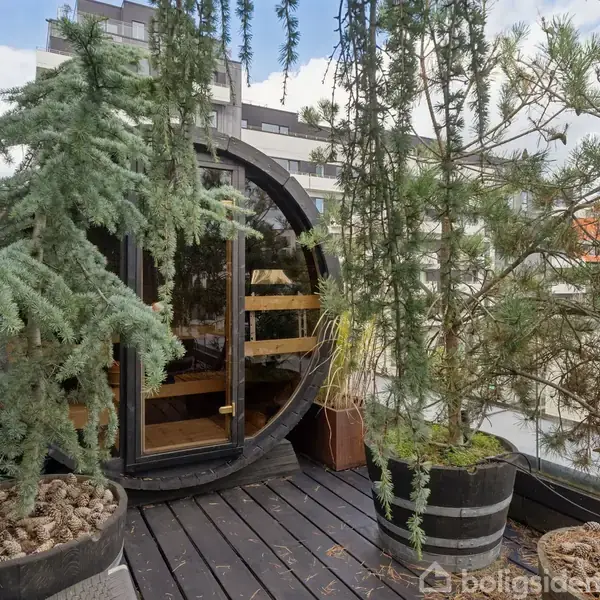 Terrassemøbler står omkring et bord med glas og flasker, beskyttet af en markise. Omgivet af grønne planter, foran en bygning med store vinduer.