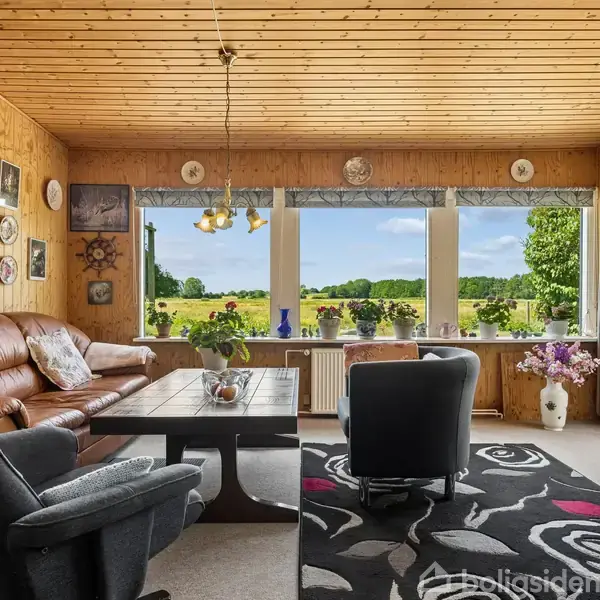 En stue med trævægge og -loft har en brun lædersofa og to lænestole omkring et sofabord. Store vinduer viser en grøn mark udenfor. Tæppet er mønstret i sort og hvidt.