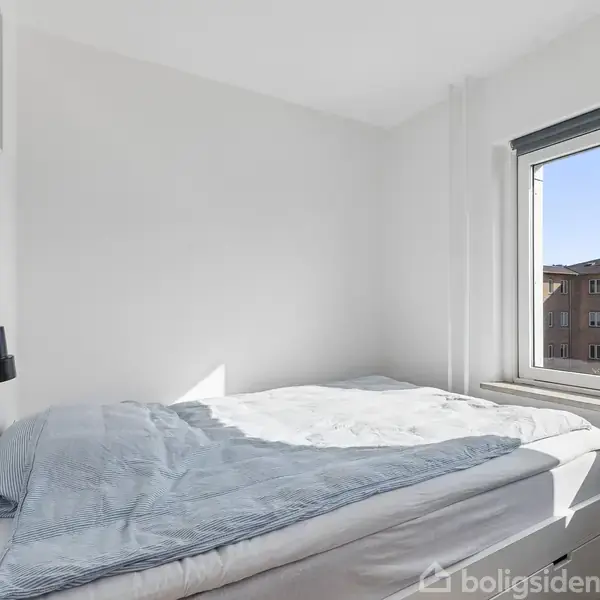 Seng med lyseblåt sengetøj står ved siden af et vindue. Udenfor ses bygninger mod en klar himmel. Værelset er hvidmalet med en enkelt væglampe monteret.