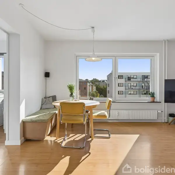 En stue med sofa mod en rød væg, et tv på stativ, og en gulvlampe. Et rundt bord med stole står ved vinduet, der viser byudsigt.
