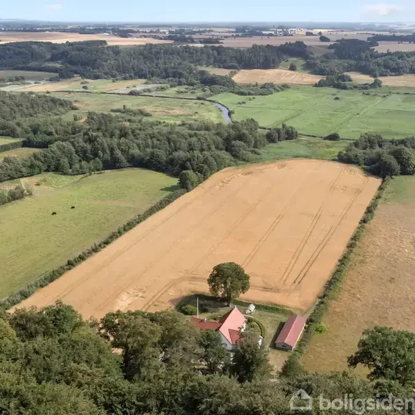 Et stort, åbent landskab med marker omgivet af skove og en bygning. Markerne er delvist høstede, og en vej løber gennem området, omgivet af frodig vegetation.