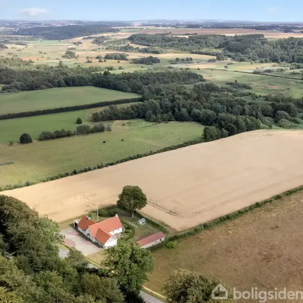 En gård med rødt tegltag beliggende i et stort, åbent markeret landskab. Omgivet af dyrkede marker og skov i det fjerne. Tæt ved en vej og spredt bebyggelse.