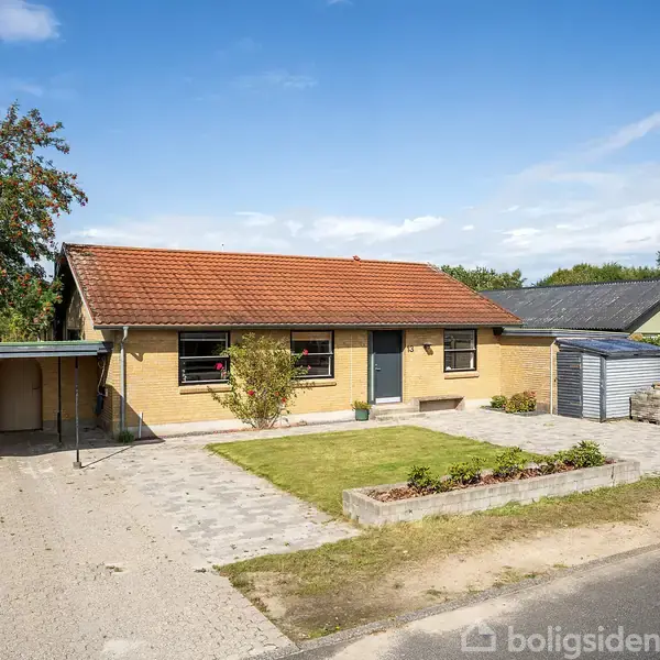 Et parcelhus med gult murværk og rødt tegltag står i solskin. Foran huset er der en lille græsplæne og en flisebelagt indkørsel. Ved siden af er et skur.