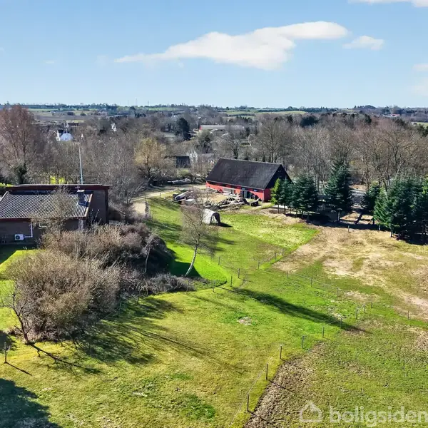 To huse står omgivet af grønt landskab og træer. Det ene hus er rødt, det andet har et mørkt tag. Omkring dem ses en græsplæne og åbne marker under en blå himmel.