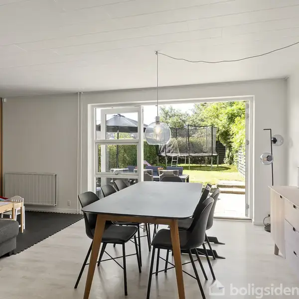 Spisebord med stole står i en lys stue. En åben dør fører ud til en have med græs og træer. Indretningen er moderne med en sort sofa og en kommode.