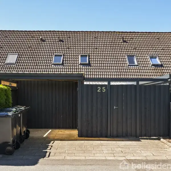 Et rækkehus med sort facade er markeret med husnummeret 25. En carport ved siden af indeholder skraldespande. Til højre står et "Til salg" skilt fra Home Portalen.