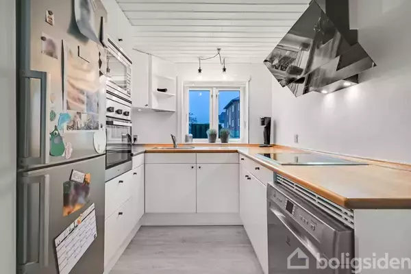 Et moderne køkken med hvide skabe og træbordplader. Køleskab, ovn og opvaskemaskine er indbygget. En bred emhætte hænger over komfuret. Vindue i baggrunden med udsigt til nabolag.