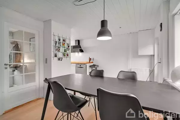 Sort spisebord med fire stole omgivet af et lyst køkkenmiljø. En pendellampe hænger over bordet, mens en glasdør og vægdekoration ses i baggrunden.