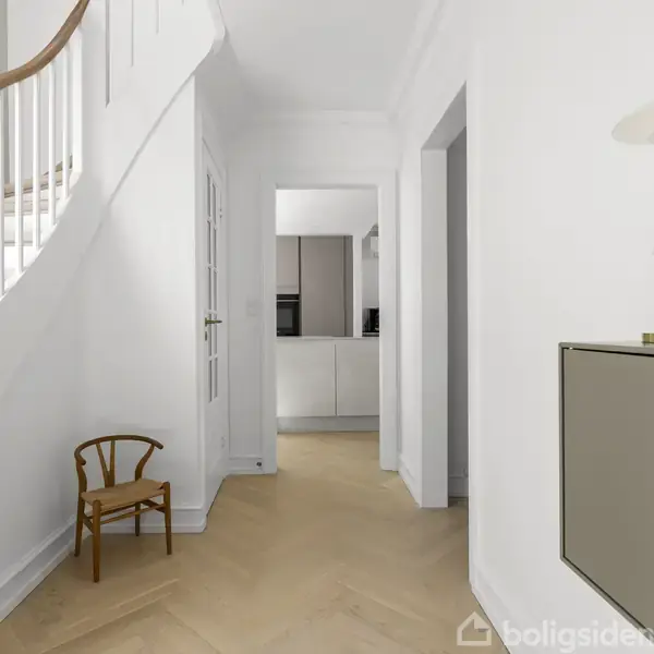 Trappe i hvidt rum med buet gelænder, lille stol til venstre. Moderne lampe og skab på højre væg. Åben dør fører til køkken.