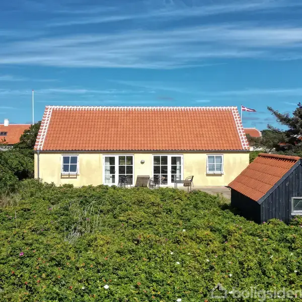 Gul bygning med rødt tegltag står stille omgivet af frodige buske. Foran ses en lille sort bygning med rødt tag, og himlen er klar med flyvende Dannebrog-flag.