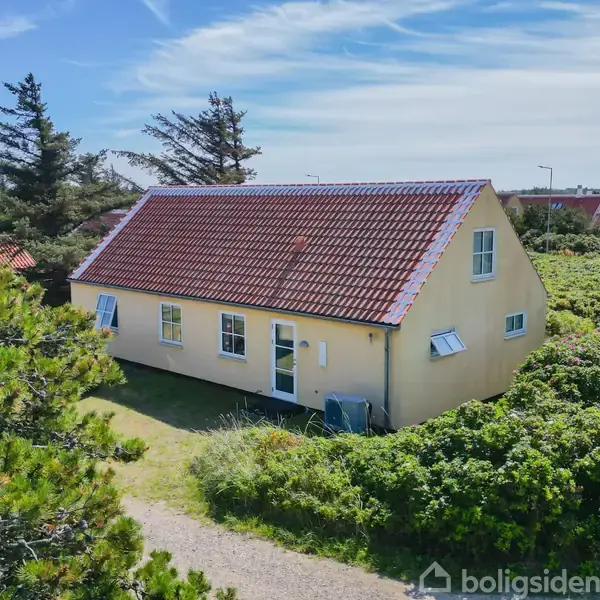 Et gult hus med rødt tegltag står stille omgivet af tæt grøn vegetation og andre huse i baggrunden under en blå himmel med lette skyer.
