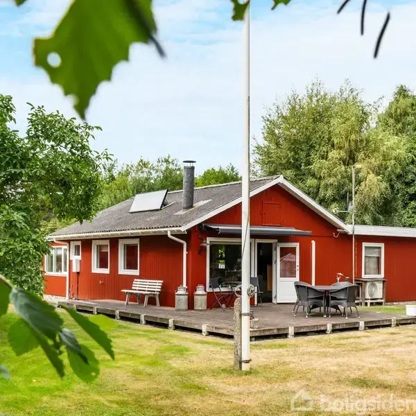 Et rødt træhus står med en terrasse og havemøbler. Omkring huset er der en grøn græsplæne og træer, der giver indtryk af en idyllisk, skovbevokset omgivelser.