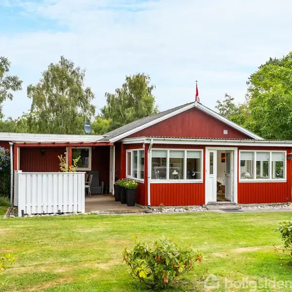 Et rødt træhus med hvidt stakit og vinduer står i en grøn have. Græs og træer omgiver huset. En carport ses til højre.