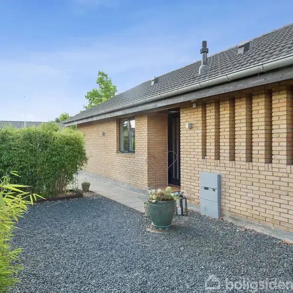 En etplans gul murstensvilla med skråt tag og flere lodrette vinduer, står omkranset af buske og en grusbelagt indkørsel. Foran huset står en grøn plante i en krukke.