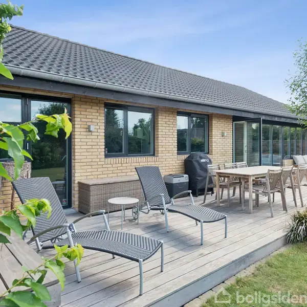 Et terrasseområde med solsenge og spisebord. Det er foran et murstenshus med store vinduer og omgivet af have. En grill står ved bordet under en klar himmel.