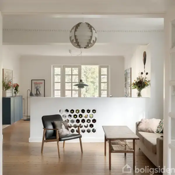 En stue med en sofa og en lænestol står foran et sofabord. I baggrunden ses en væg med dekorative åbninger, og rummet er lyst op af naturligt lys fra vinduerne.