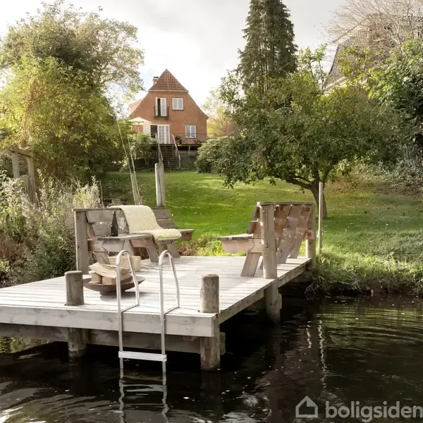 En træbrygge med bænke og en badestige står ved en sø. Baggrunden viser en græsplæne med et stort gammelt hus omgivet af træer og buske.