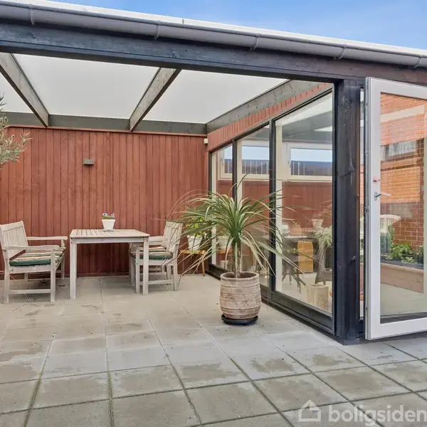 En møbleret terrasse med havemøbler og planter. Terrassen er overdækket med glas og omgivet af røde trævægge og en murstensvæg. En glasdør står åben mod en delvist synlig indendørs plads.