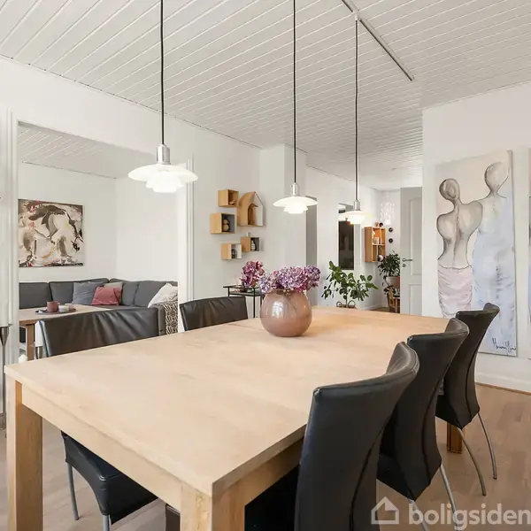Et spisebord med sorte stole står i en moderne spisestue med lyse vægge. Der hænger lamper over bordet, og der står en vase med lilla blomster. En reol med glas står ved siden af.