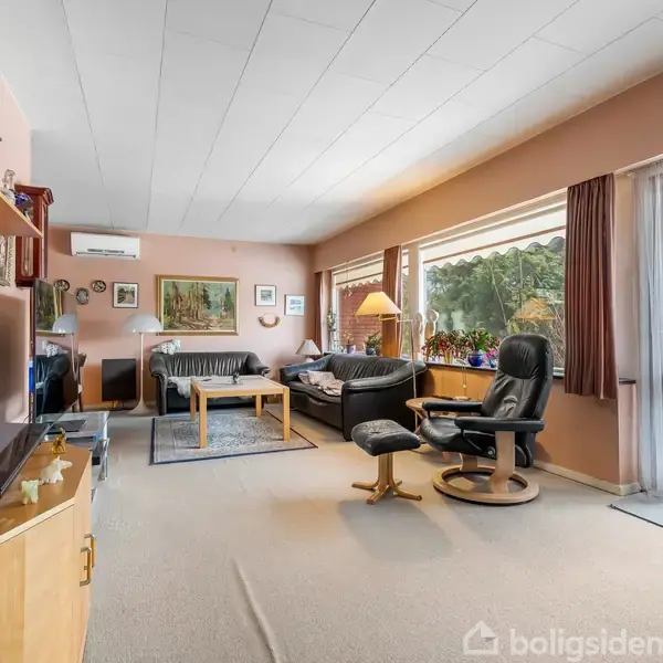 Stue med sofagruppe og lænestol ved et stort vindue. Reoler fyldt med bøger og dekorative genstande omkranser rummet. Belysning fra en gulvlampe skaber en hyggelig atmosfære.