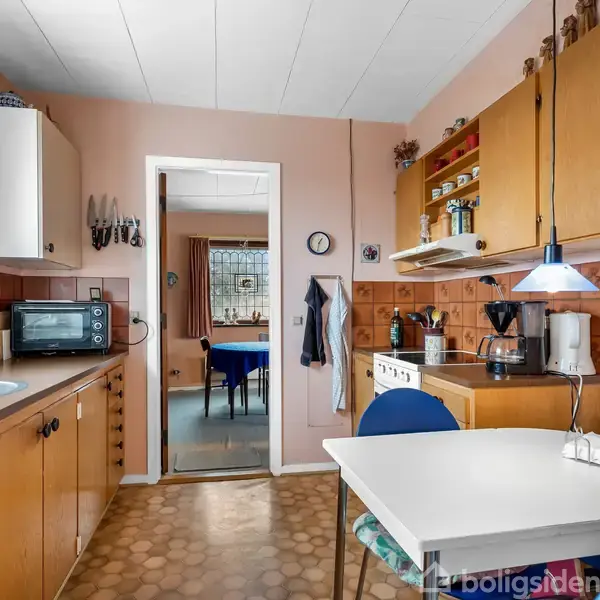 Et køkken med et hvidt bord og blå stole, en mikrobølgeovn på køkkenbordet til venstre. En døråbning fører til en stue. Flere skabshylder og køkkenapparater.