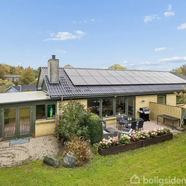 Huset har solpaneler på taget, store vinduer og en terrasse med havemøbler. Det er omgivet af en grøn have med buske og træer i en fredelig boligområde.