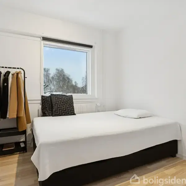 En enkeltseng med hvidt sengetøj står i et lyst soveværelse. Ved siden af er et tøjstativ med jakker. Et vindue med udsigt viser træer udenfor.