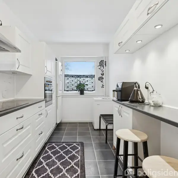 Et moderne køkken med hvide skabe og sorte bordplader. Køkkenet har en ovn og komfur, en opvaskemaskine, samt en lang bar med to taburetter. Et vindue med planter skaber lysindfald.