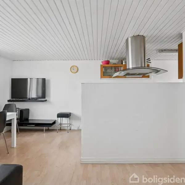 Værelse med sort fjernsyn hængende på en hvid væg ved siden af et bord med stole; et køkkenområde med emhætte. Trægulv og hvidt loft under naturligt lys fra vindue.