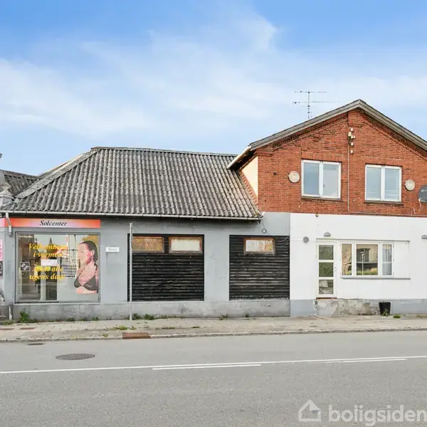 Bygning med en rød murstensfacade ligger ved vejkryds. Til venstre er en frisørsalon med skiltet "Salonaux" og billede af en kvinde. Vejret er klart med blå himmel.