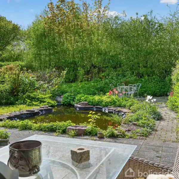 Terrassebord med stole står ved en glasbord, hvorpå der er en urtepotte. En lille dam med stenkanter ses i en frodig have med grønne buske og træer. I baggrunden er der et bord med stole.