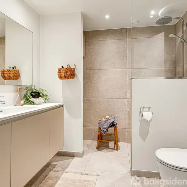 Toilet står stille i moderne badeværelse med lyse fliser. Til venstre er der en vask med spejl og skab, samt en plante. Brusebad til højre. Vasketøjsstol i midten.