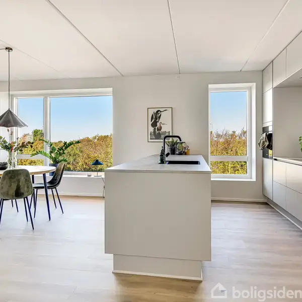 Et lyst køkken med et stort køkkenbord og indbyggede skabe. Et spisebord med stole ved siden af store vinduer, der giver udsigt til grønne træer udenfor.