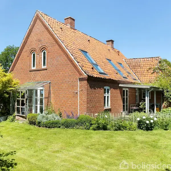 Rødt murstenshus med skråt tegltag og ovenlysvinduer står omgivet af frodig have med velplejede græsplæner og buske. En lille veranda ses til venstre.