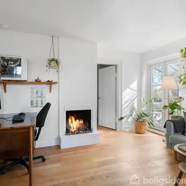 Et hyggeligt stueområde med en tændt pejs, et skrivebord med stol til venstre, grønne planter, en sofa til højre og et bord med bøger. Store vinduer lader naturligt lys ind.