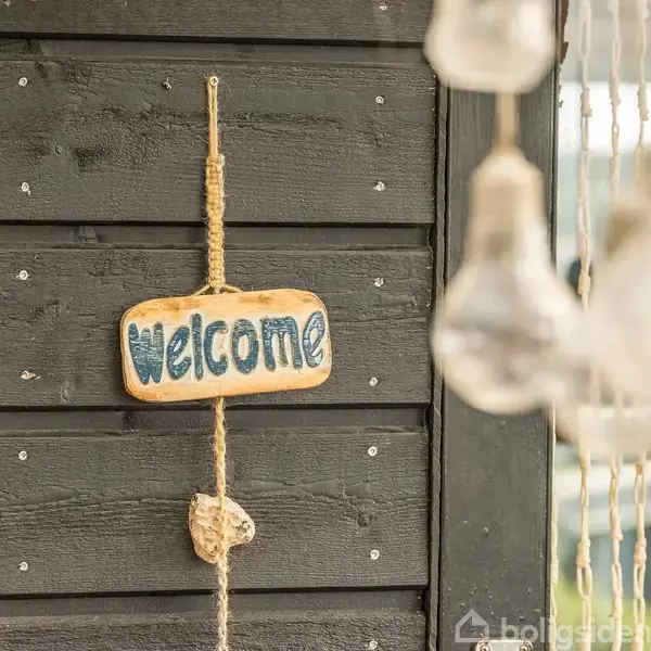Et træskilt med teksten "welcome" hænger på en sort trævæg. I forgrunden hænger dekorative pærer, hvilket skaber en varm og indbydende atmosfære.