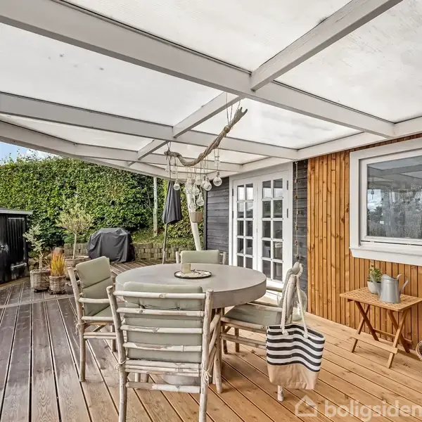 Terrasseområde med rundt bord og seks stole under en gennemsigtig overdækning. Ved siden af står en plante og små møbler. Omgivet af trævægge og grønne buske.